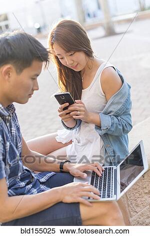 Stock Photography - Korean couple using laptop and cell phone outdoors. Fotosearch