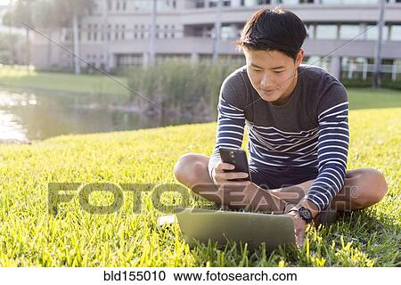 Stock Image - Korean man using laptop and cell phone in grass. Fotosearch