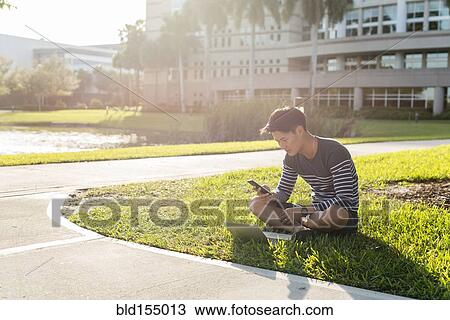 Stock Image - Korean man using laptop and cell phone in grass. Fotosearch