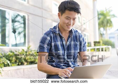 Stock Image - Korean man using laptop outside building. Fotosearch