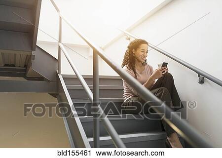 Stock Image - Low angle view of mixed race businesswoman using cell phone on staircase. Fotosearch