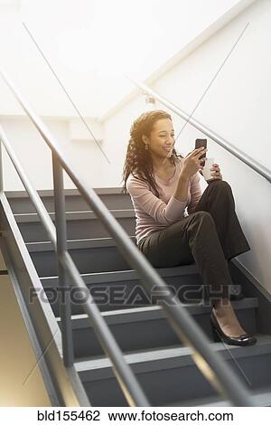Stock Image - Low angle view of mixed race businesswoman using cell phone on staircase. Fotosearch