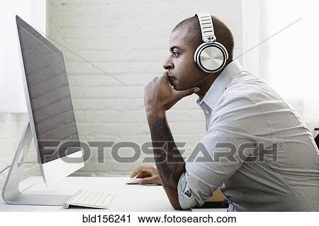 Stock Image - Mixed race businessman listening to headphones and working at desk. Fotosearch