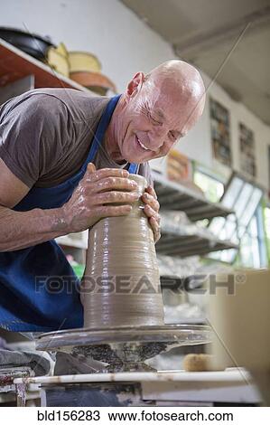 Stock Image - Older Caucasian man forming pottery on wheel in ceramics studio. Fotosearch