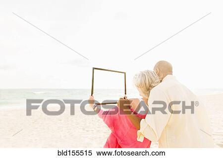 Stock Photography - Older couple holding empty picture frame on beach. Fotosearch