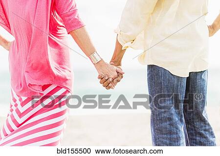 Stock Image - Older couple holding hands on beach. Fotosearch