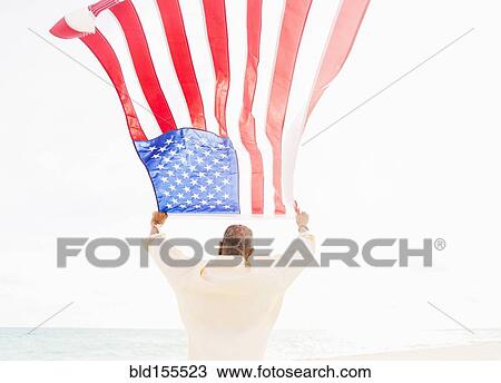 Stock Image - Older mixed race man holding American flag on beach. Fotosearch