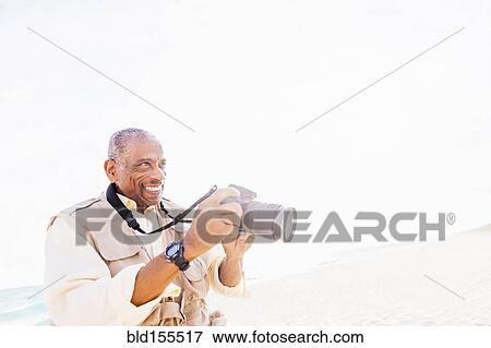 Stock Photo - Older mixed race man photographing on beach. Fotosearch