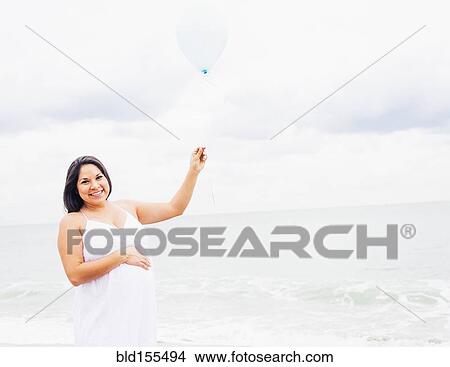 Picture - Pregnant mixed race woman holding balloon on beach. Fotosearch
