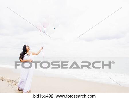 Stock Image - Pregnant mixed race woman holding balloons on beach. Fotosearch
