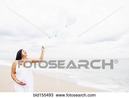Stock Image - Pregnant mixed race woman holding balloons on beach. Fotosearch