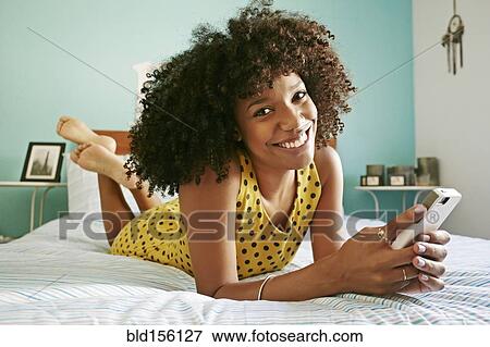 Stock Photo - Smiling mixed race woman using cell phone on bed. Fotosearch