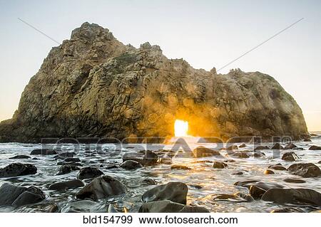 Stock Photo - "Sun shining through rock formation to ocean waves, Big Sur, California, United States". Fotosearch