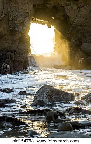 Stock Photo - Sunbeam shining through rock formation to ocean waves. Fotosearch