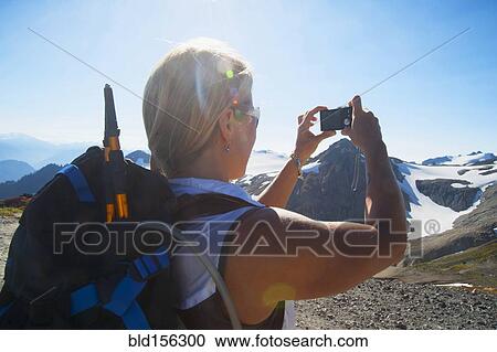 Stock Image - Woman taking photograph of remote mountains. Fotosearch
