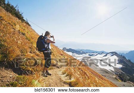 Stock Image - Woman taking photograph of remote mountains on hillside. Fotosearch