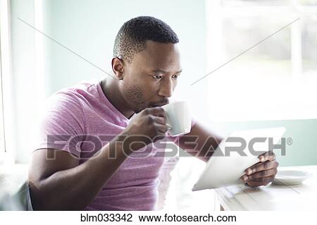 Stock Image - Black man drinking coffee using digital tablet. Fotosearch
