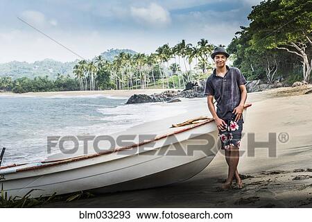 Stock Image - Hispanic man standing near boat on beach. Fotosearch
