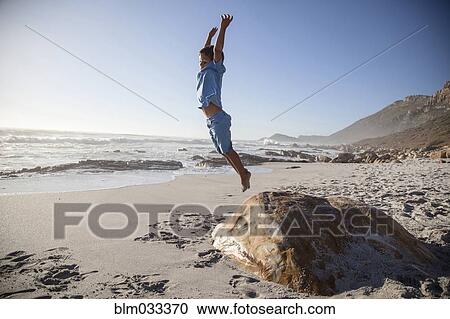 Stock Image - Mixed race boy jumping off rock on beach. Fotosearch