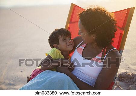 Stock Image - Mixed race mother and daughter hugging in chair on beach. Fotosearch
