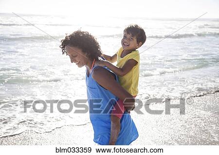 Stock Photo - Mixed race mother carrying daughter piggyback on beach. Fotosearch