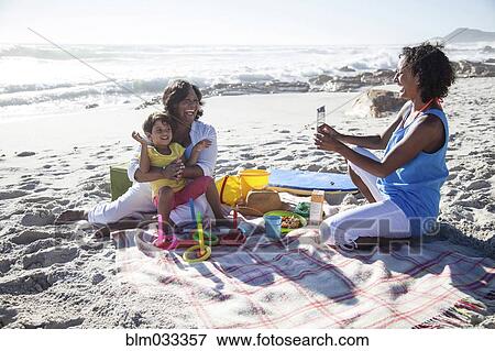 Stock Photo - Mixed race mother photographing grandmother and daughter at beach. Fotosearch