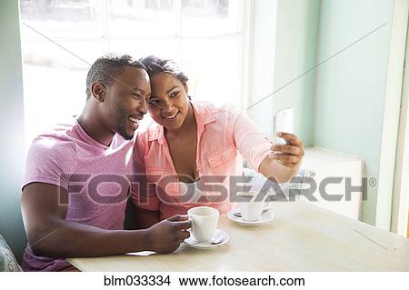Picture - Smiling couple taking cell phone selfie at table with coffee. Fotosearch