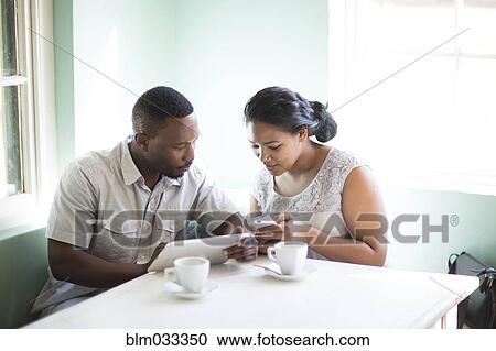 Stock Image - Smiling couple using cell phone and digital tablet at breakfast. Fotosearch