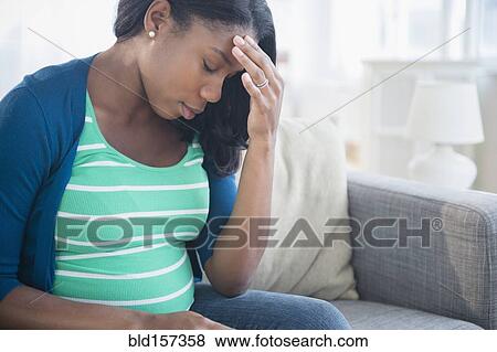 Stock Photo - Anxious Black pregnant woman rubbing forehead on sofa. Fotosearch
