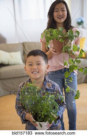 Stock Image - Asian brother and sister holding potted plants in living room. Fotosearch