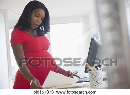 Stock Image - Black pregnant businesswoman reading paperwork at desk in office. Fotosearch