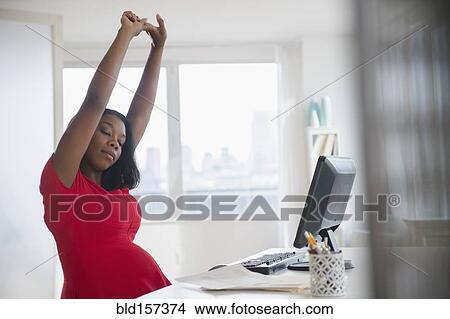 Picture - Black pregnant businesswoman stretching arms at desk in office. Fotosearch