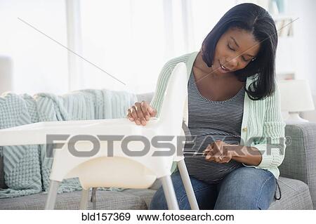 Stock Photo - Black pregnant woman assembling high chair in living room. Fotosearch
