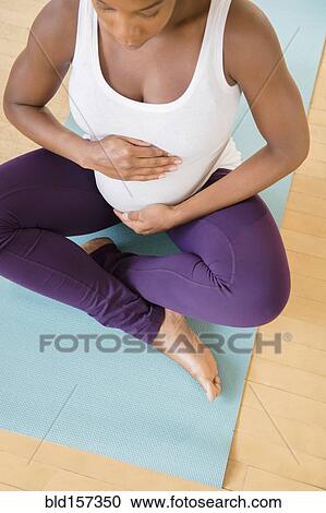Stock Image - Black pregnant woman sitting on exercise mat holding stomach. Fotosearch
