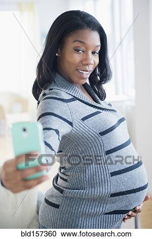 Stock Image - Black pregnant woman taking cell phone selfie. Fotosearch