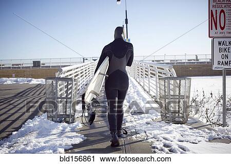 Stock Image - Black teenage girl in wetsuit carrying surfboard in winter. Fotosearch