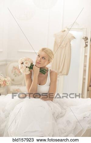 Stock Image - Bride sitting with bouquet on bed thinking. Fotosearch