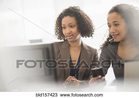 Stock Image - Businesswomen working with cell phone and computer in office. Fotosearch