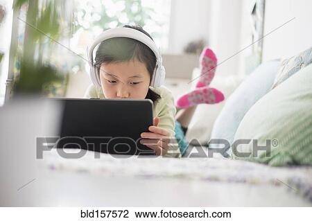 Stock Image - Chinese girl using digital tablet with headphones on bed. Fotosearch