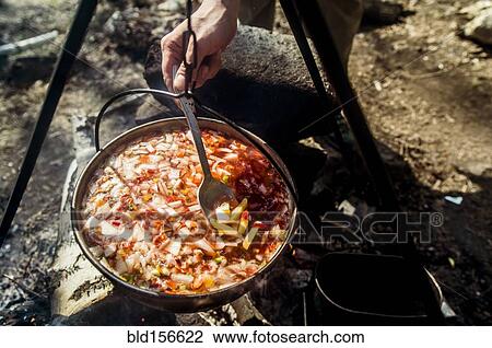 Stock Image - Close up of person stirring soup cooking over campfire. Fotosearch