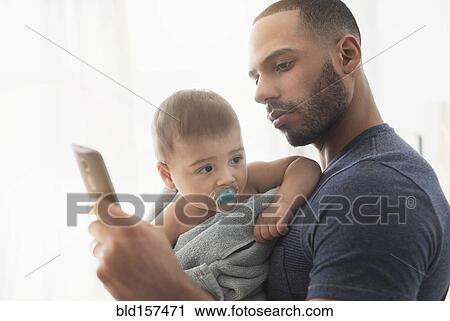 Stock Image - Father holding baby son and using cell phone. Fotosearch