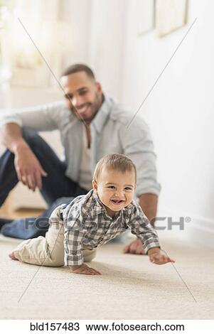 Stock Image - Father watching baby son crawling on floor. Fotosearch