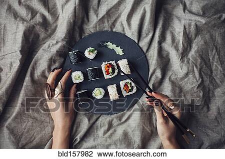 Stock Image - Hands of woman reaching for platter of sushi on bed. Fotosearch