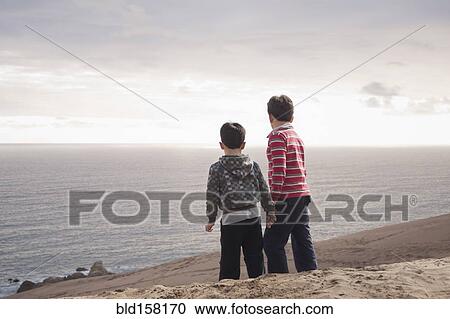 Stock Image - Hispanic brothers standing on sand dunes on beach. Fotosearch
