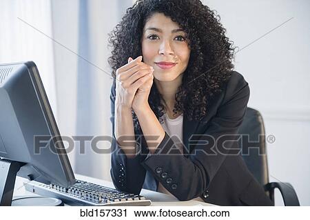 Stock Image - Mixed race businesswoman smiling at desk in office. Fotosearch