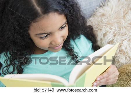 Stock Photo - Mixed race girl reading book on sofa. Fotosearch