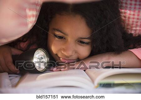 Stock Image - Mixed race girl reading book with flashlight under blanket. Fotosearch