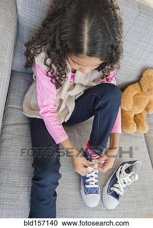 Stock Image - Mixed race girl tying her shoes on sofa. Fotosearch