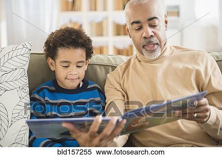 Stock Photography - Mixed race grandfather and grandson reading book on sofa. Fotosearch