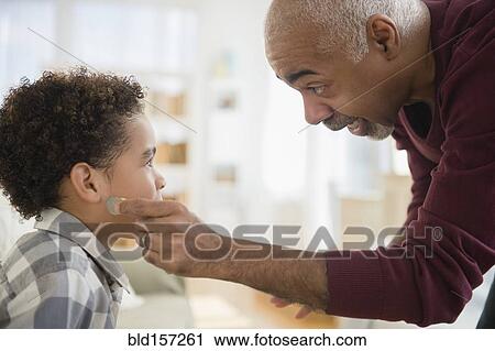 Stock Image - Mixed race grandfather pulling magic coin from ear of grandson. Fotosearch